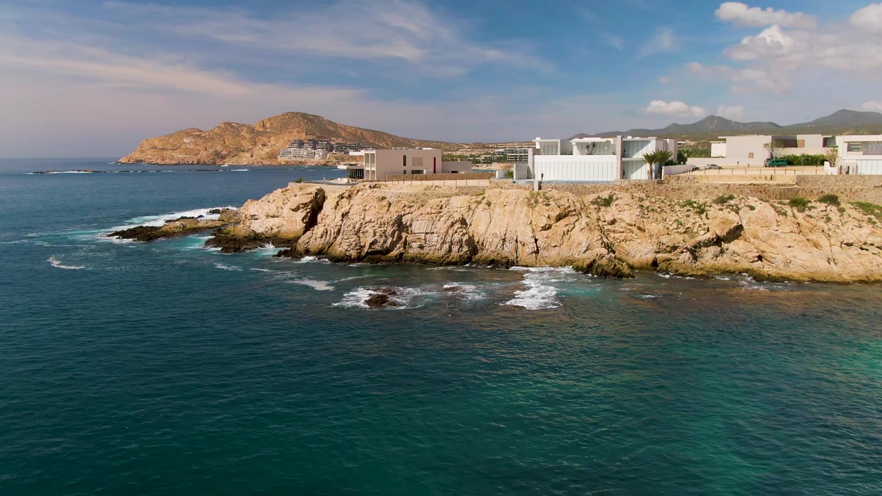 At 0:02, the El Tule Los Cabos shoreline remains centered, with darker blue water in front, foam curling around dark rocks, modern coastal buildings above the cliff, hills beyond, and no visible people.