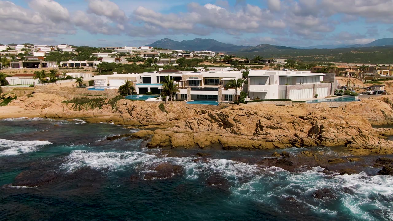 Opening aerial frame in El Tule Los Cabos shows white modern buildings on tan rocks above dark blue surf, with distant mountains under broken clouds and no visible people.