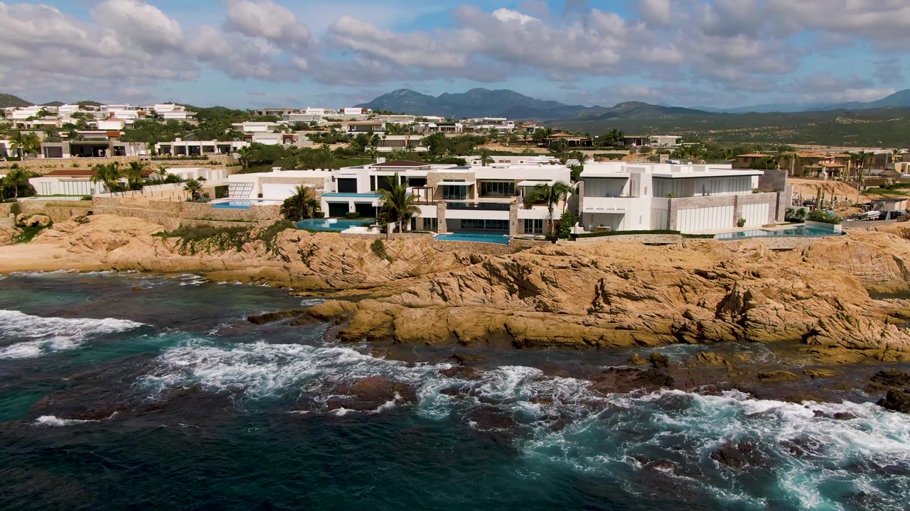 At 0:01 in El Tule Los Cabos, foamy waves spread farther across the foreground rocks below the white coastal buildings, with mountains and clouds still visible and no visible people.