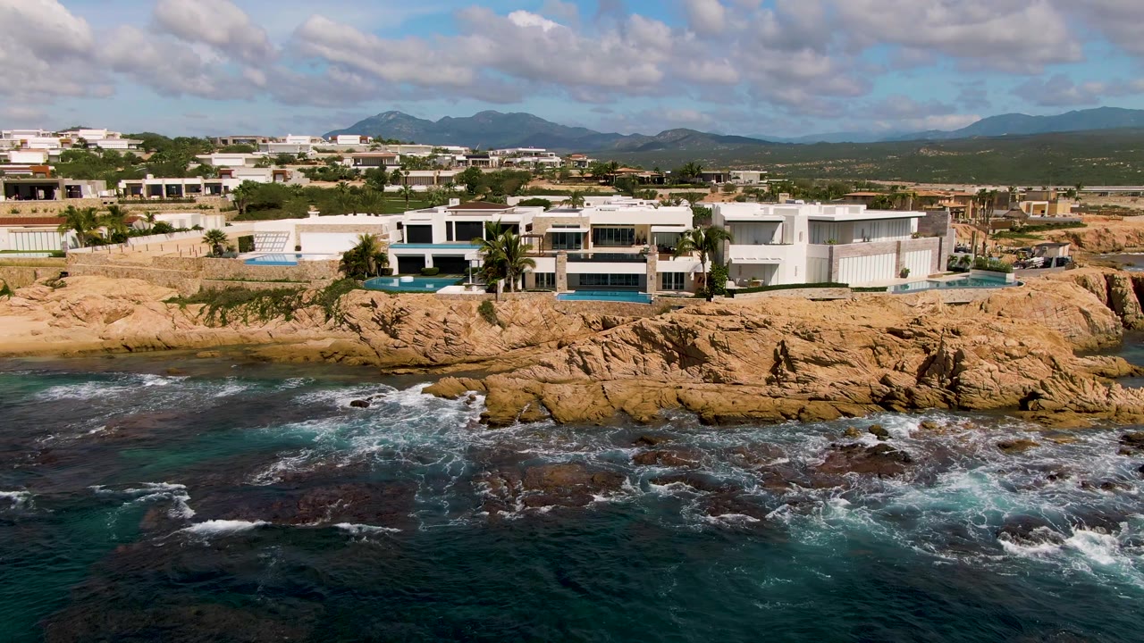 Aerial view in El Tule Los Cabos, Mexico, with modern white buildings above tan rocky cliffs, dark blue ocean in front, distant mountains, scattered clouds, and no visible people.