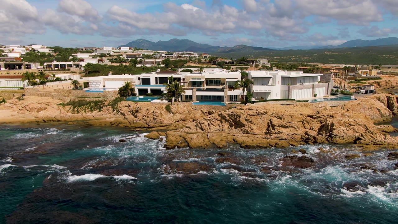 Aerial frame from El Tule Los Cabos with white cliffside buildings centered over rocky shore, foamy surf spreading across the lower rocks, mountains behind, and no visible people.