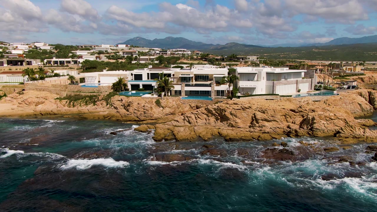 El Tule Los Cabos aerial view with white coastal buildings and visible pools above ocher rocks, waves curling along the shoreline, broad sky, and no visible people.