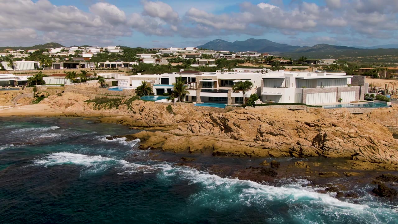 Modern white homes in El Tule Los Cabos sit above tan rocks as dark blue water breaks across the lower foreground, with distant mountains and no visible people.