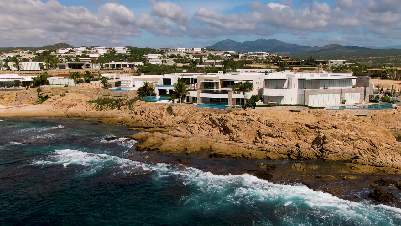 White oceanfront homes, pools, rocky ledge, and surf under clouds.