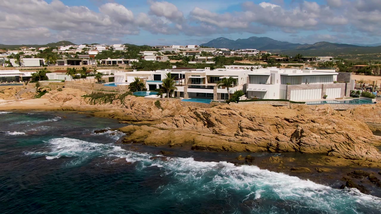 An aerial view in El Tule Los Cabos shows white homes spanning the midground as waves push white foam against the rocky shoreline, with no visible people.