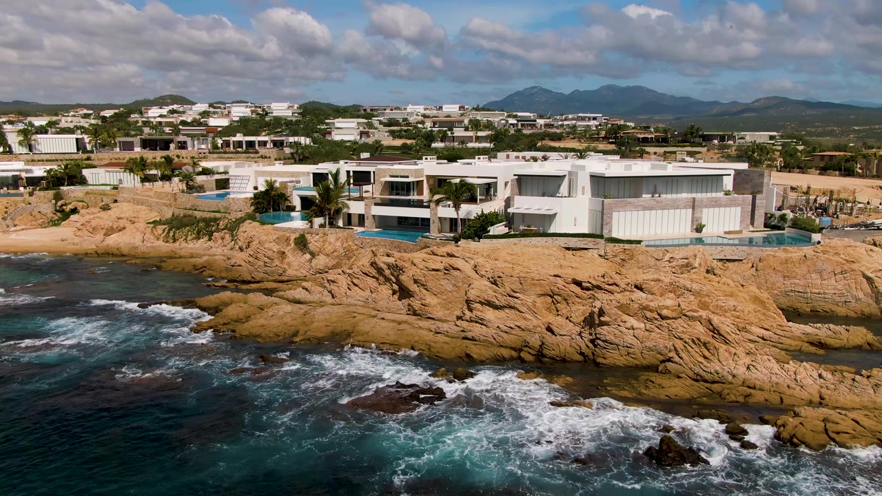 Aerial frame from El Tule Los Cabos showing white modern homes above tan rocky shoreline, dark blue water, breaking surf, scattered clouds, distant hills, and no visible people.