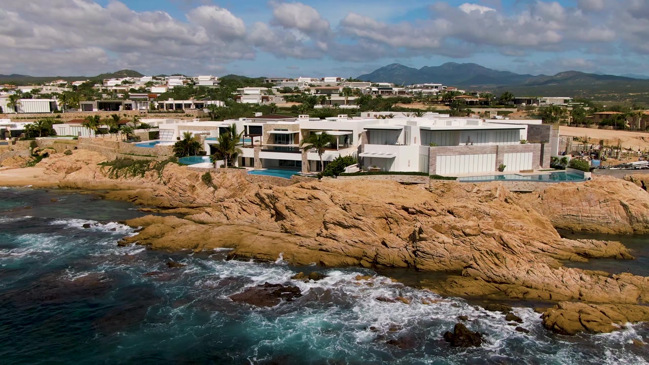 At 0:01 in El Tule Los Cabos, surf spreads over dark rocks below white coastal buildings and a low hillside neighborhood, with no visible people in the frame.