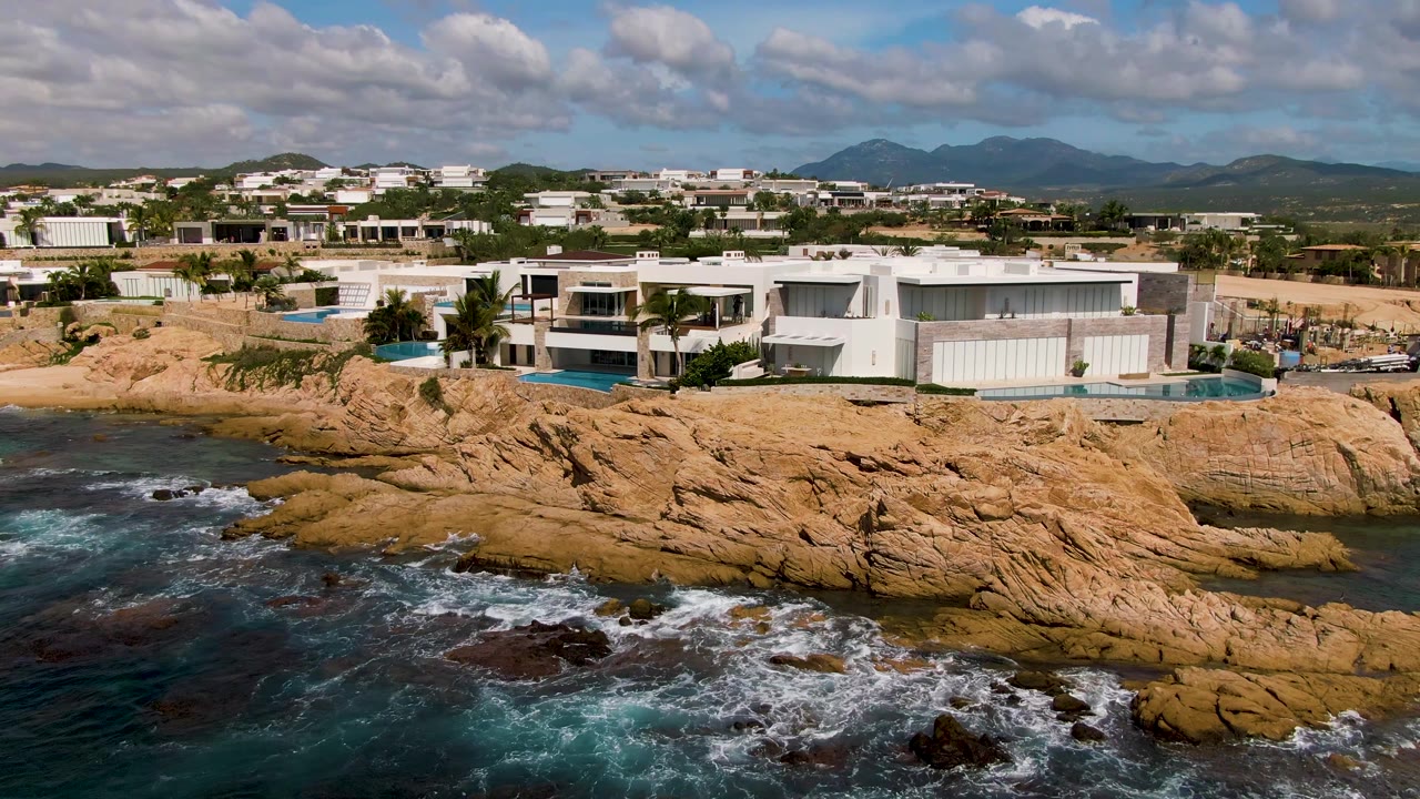 Aerial view in El Tule Los Cabos with a white rectangular home beside a pool, tan coastal rocks, churning blue water, cloud shadows, distant hills, and no visible people.