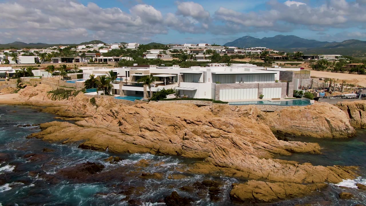 Wide elevated view of El Tule Los Cabos, Mexico, with tan rock formations, blue-green surf, modern shorefront buildings, distant mountains, scattered clouds, and no visible people.