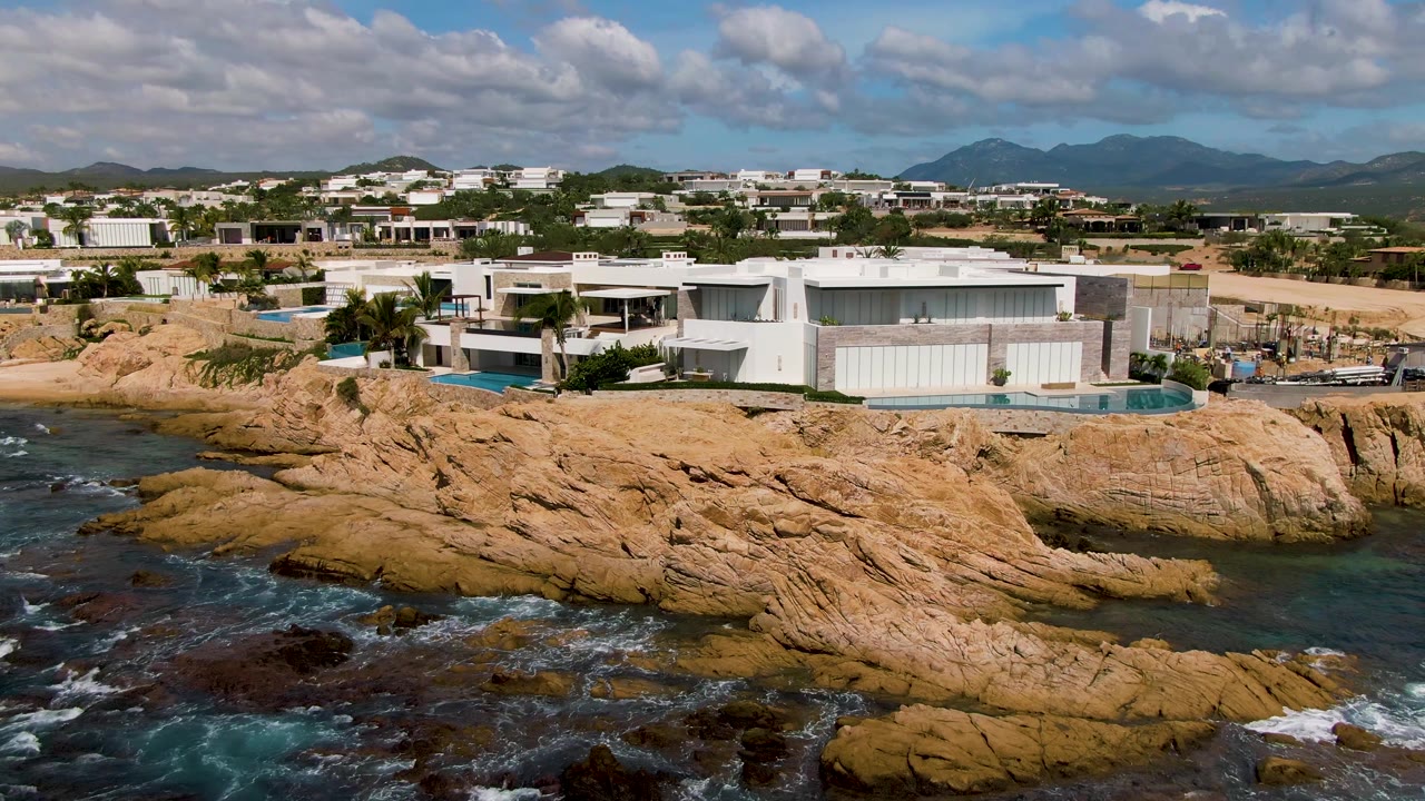El Tule Los Cabos coastline with white modern villas above layered tan rocks, darker reef areas in the water, small patches of white surf, mountains, clouds, and no visible people.