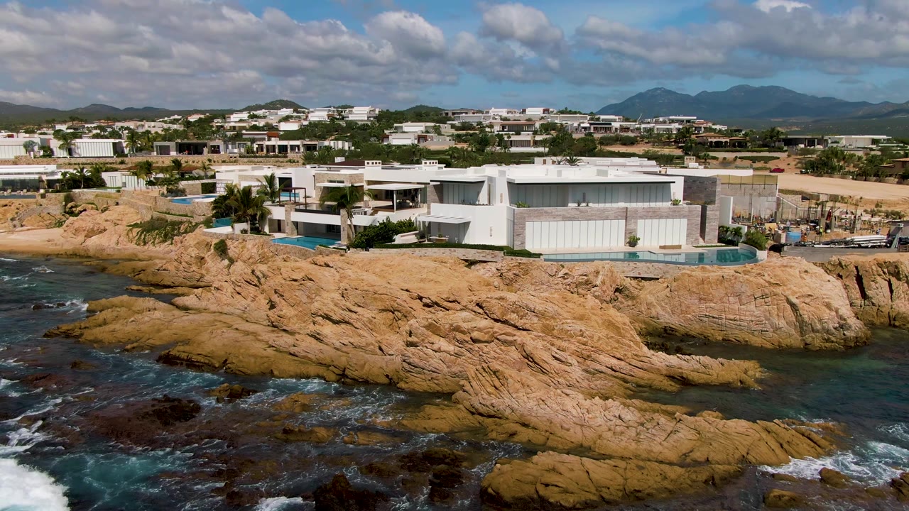 Wide coastal frame in El Tule Los Cabos, Mexico, showing modern white buildings on the shore, tan rocks across the foreground, blue-green water, breaking white foam near the lower edge, and no visible people.