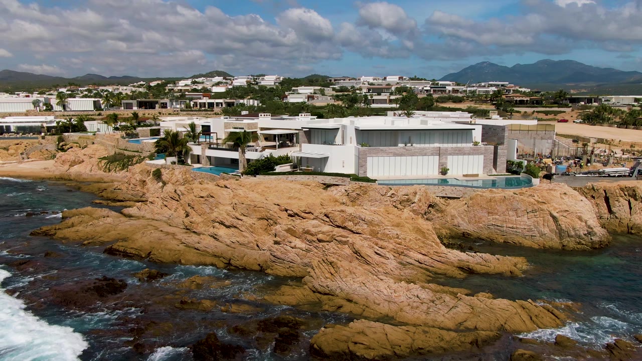 Ocean waves push into rocks below modern buildings and a partly cloudy sky.
