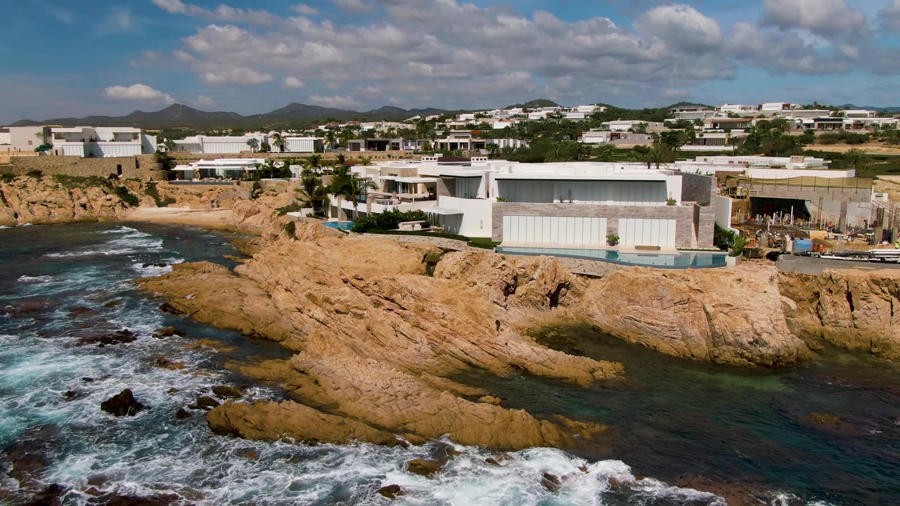 Aerial view of El Tule Los Cabos rocky coastline, with white surf at the lower left, tan rocks across the foreground, modern buildings on the cliff, and no visible people.