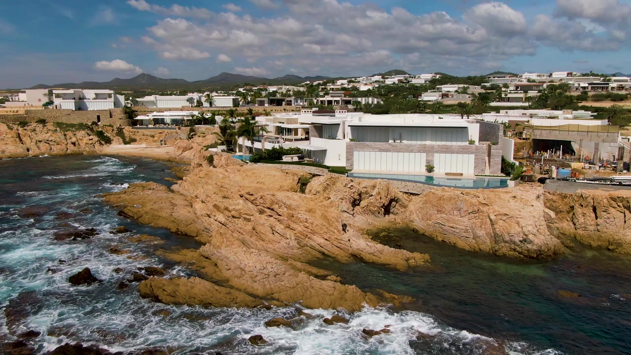 Waves break beside tan coastal rocks and cliffside buildings.