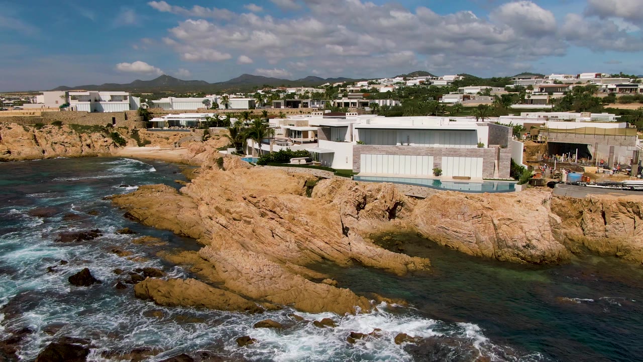 Aerial frame of El Tule Los Cabos with surf curling over the rocky shore, a pale modern building centered above the cliff, and no visible people.