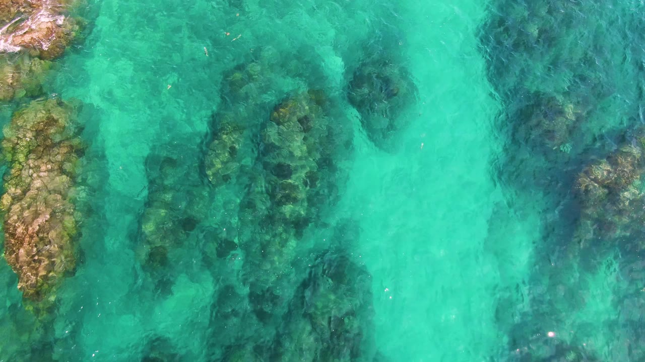 Overhead view of clear turquoise water at El Tule Los Cabos with dark submerged rock patches and no visible people.