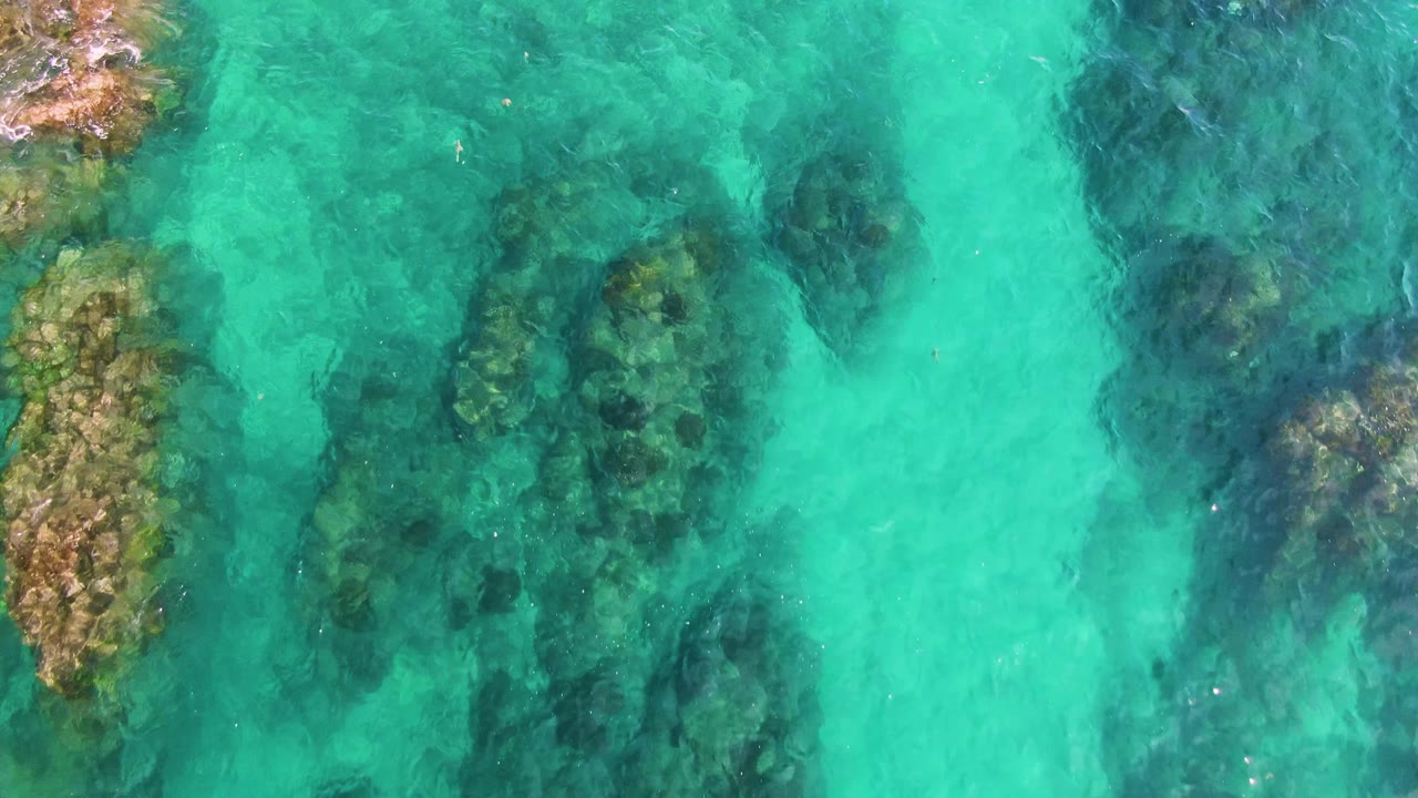 Clear green-blue water at El Tule Los Cabos reveals clustered underwater rocks near the center and no visible people.