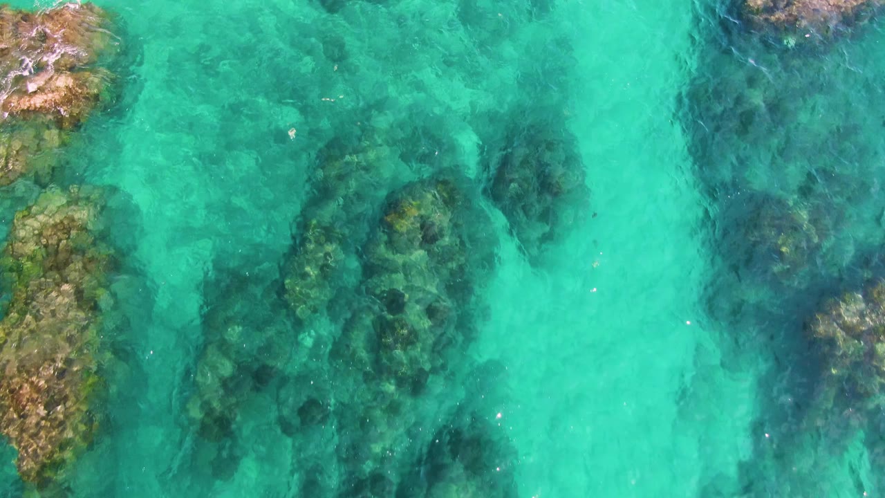 Turquoise ocean water at El Tule Los Cabos shows uneven submerged rocks and darker patches through the transparent surface with no visible people.