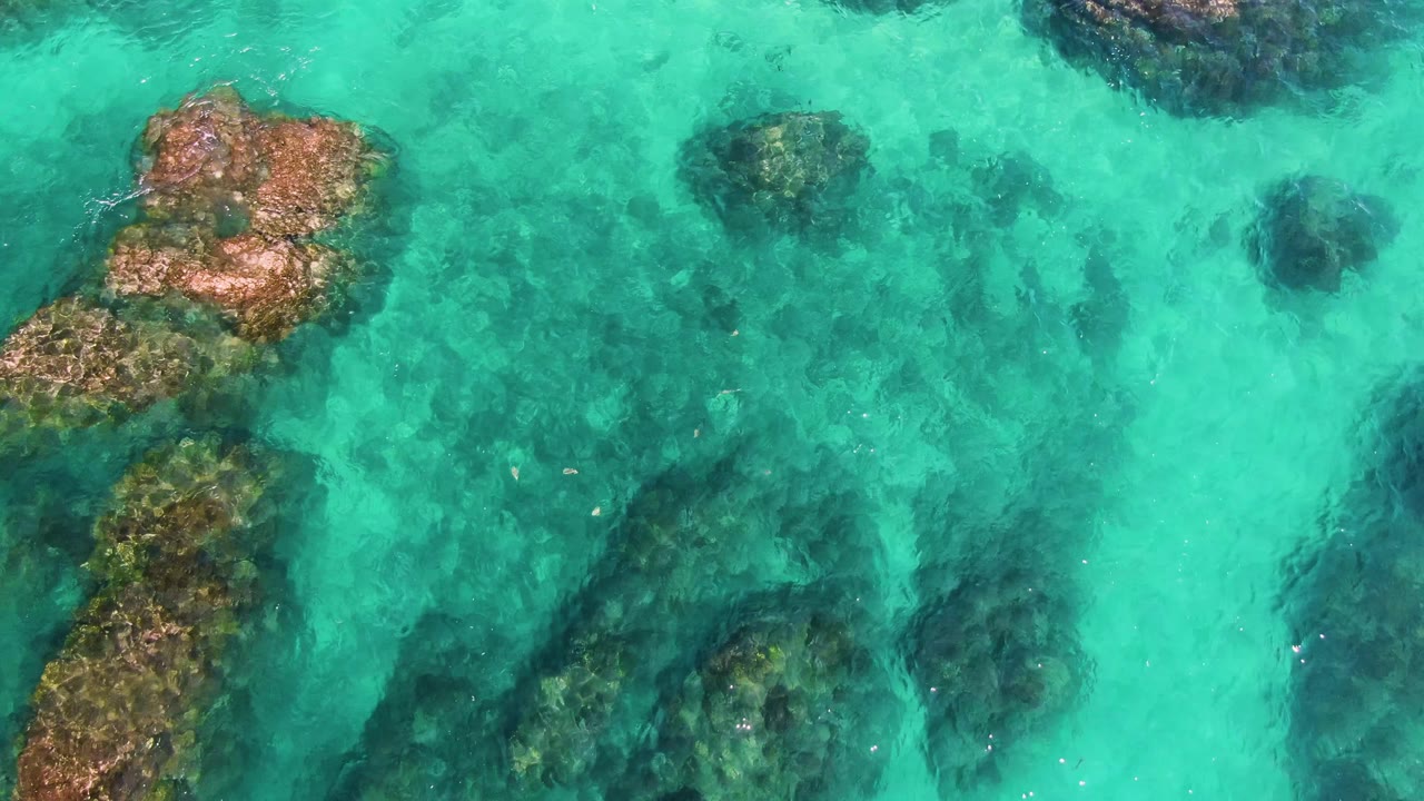Turquoise water at El Tule Los Cabos covers dark reef-like rocks along the left and lower portions of the frame, with a brighter open channel through the middle, no visible people.