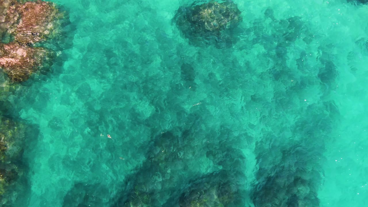 Aerial view of turquoise water at El Tule Los Cabos with broad submerged rock shadows and a warmer rocky edge at upper left, no visible people.