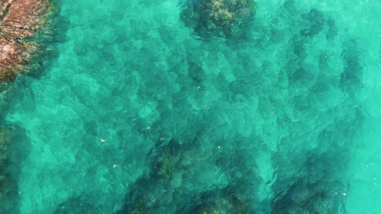 Turquoise ocean water at El Tule Los Cabos covers a mottled rocky seabed with darker formations in the lower half and a partial rock edge at left, no visible people.