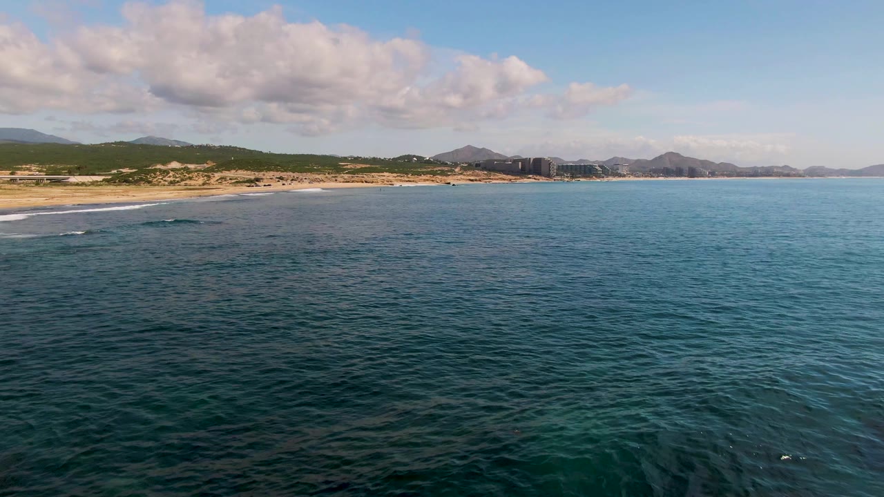 At El Tule Los Cabos, blue-green ocean fills the foreground while a sandy beach, low hills, distant mountains, scattered clouds, and no visible people appear beyond it.