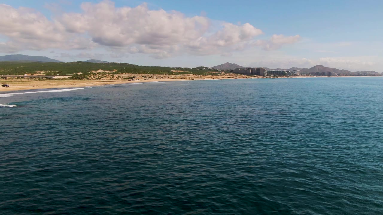 At 0:02 in El Tule Los Cabos, the calm ocean extends across most of the frame with the sandy shoreline at left, low coastal vegetation, distant mountains, soft clouds, and no visible people.