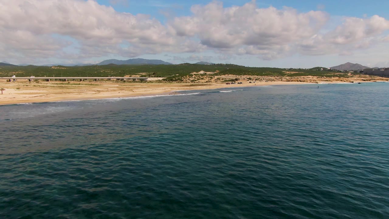Aerial frame of El Tule Los Cabos with blue-green ocean in the foreground, a pale sandy beach beyond it, distant green hills under clouds, and no visible people.