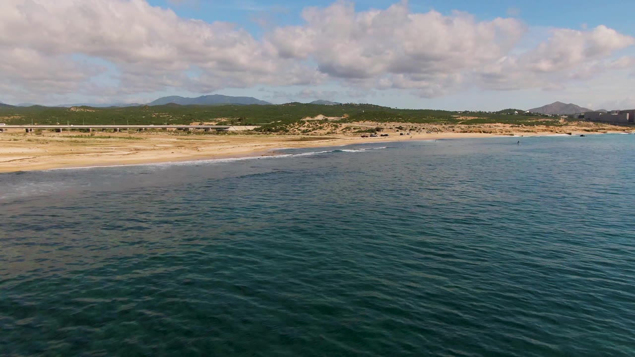 Ocean surface and sandy shoreline with low dunes and hills in the distance.