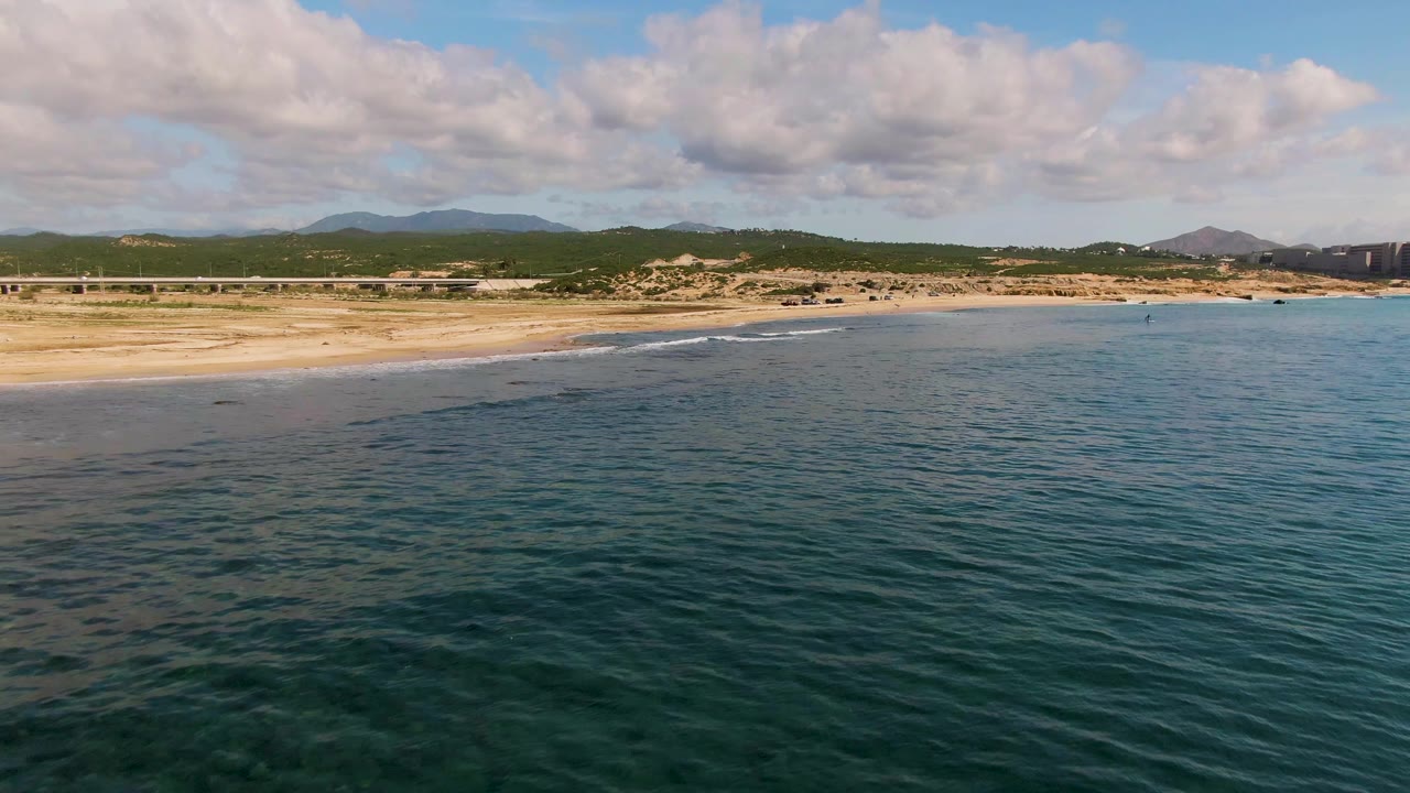 Wide aerial frame of El Tule Los Cabos showing dark blue-green water, a sandy beach, low hills, a cloud-filled sky, distant buildings at the right edge, and no visible people.