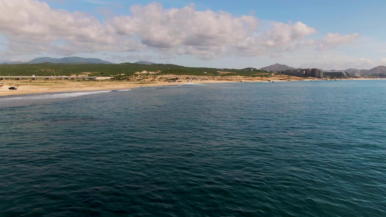Opening frame of El Tule Los Cabos with blue-green ocean in front, sandy beach along the left shore, distant hills under scattered clouds, and no visible people.