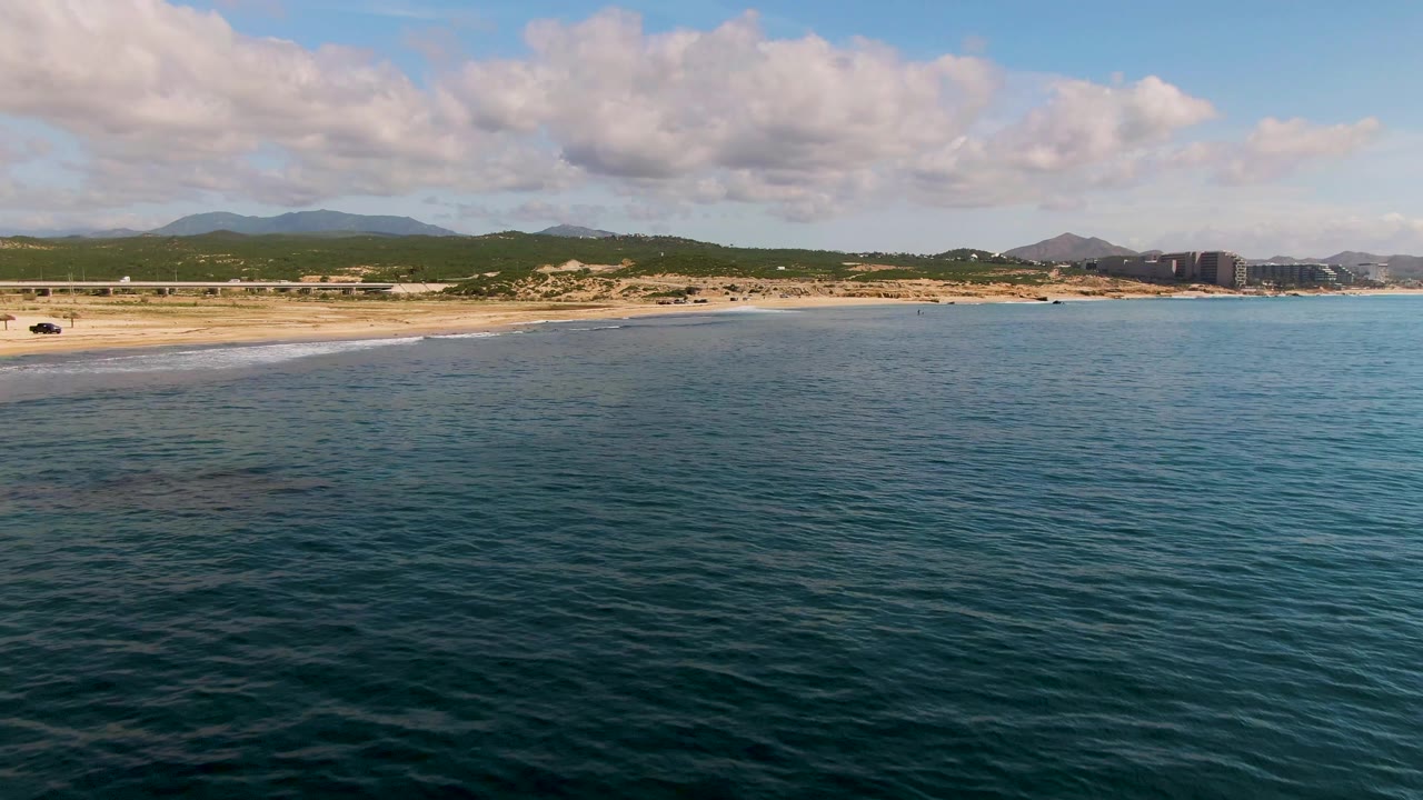 At one second, the El Tule Los Cabos shoreline stretches across the frame with rippled ocean in the foreground, a small vehicle near the sand, distant hills, and no visible people.