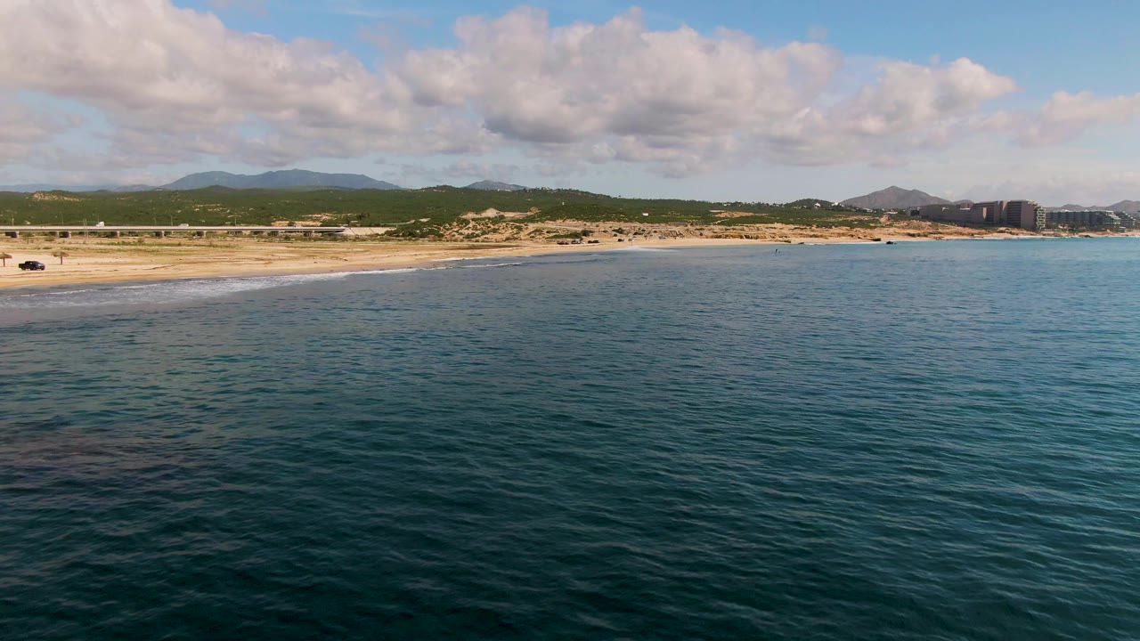 At two seconds, the El Tule Los Cabos coast remains in a wide aerial composition, with darker ocean texture in front, pale sand at left, hills and clouds beyond, and no visible people.