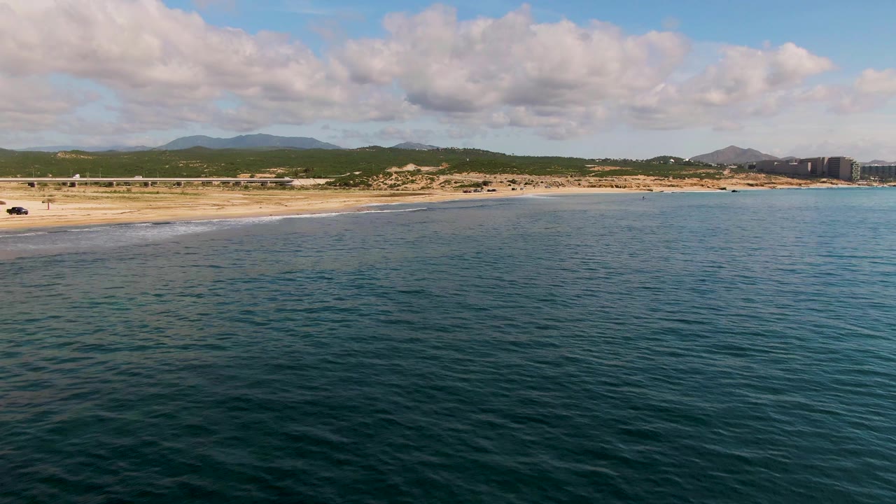 Wide beach and ocean aerial with shoreline vehicle, surf, hills, and cloud cover.