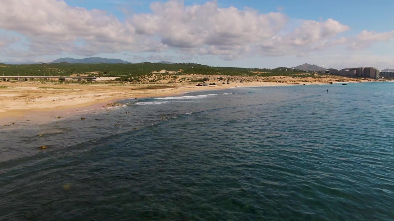 Aerial view of waves approaching a sandy beach with hills and clouds beyond.