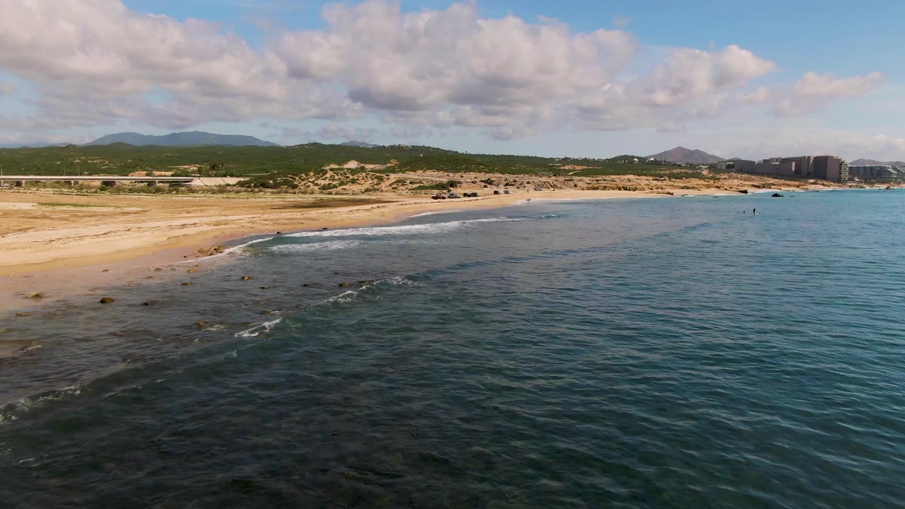 Aerial view at 0:01 over El Tule Los Cabos, Mexico, as a thin wave line reaches the sandy shore with hills, distant buildings, and no visible people.