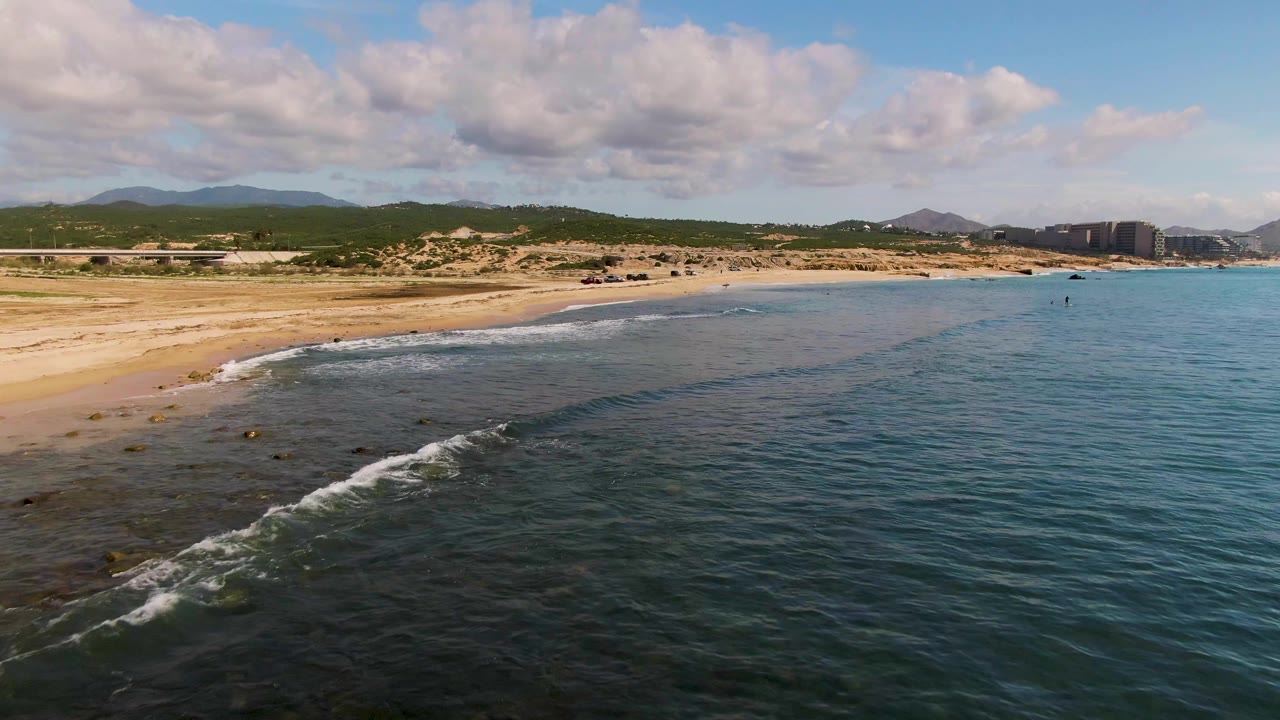At 0:02 in El Tule Los Cabos, Mexico, a longer white foam line runs diagonally across shallow water toward the beach, with hills, buildings, and no visible people.