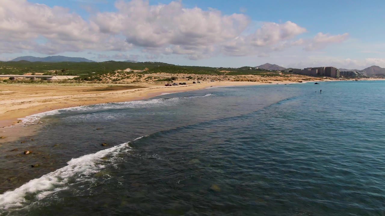 Mid-clip aerial frame over El Tule Los Cabos, Mexico, with white foam spreading in the lower-left foreground, the beach curving inland, and no visible people.