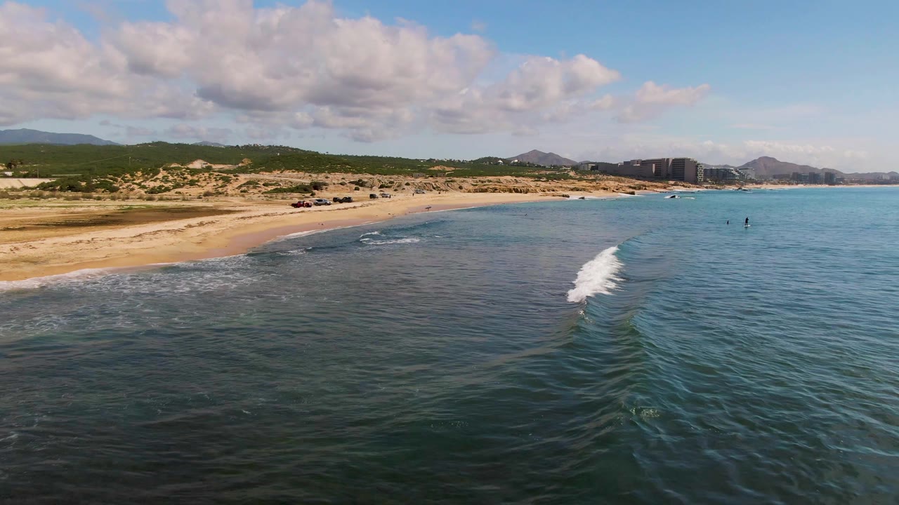 Aerial view over El Tule Los Cabos with blue-green water, a narrow white wave line, sandy beach, dunes, distant hills, scattered clouds, and no visible people.