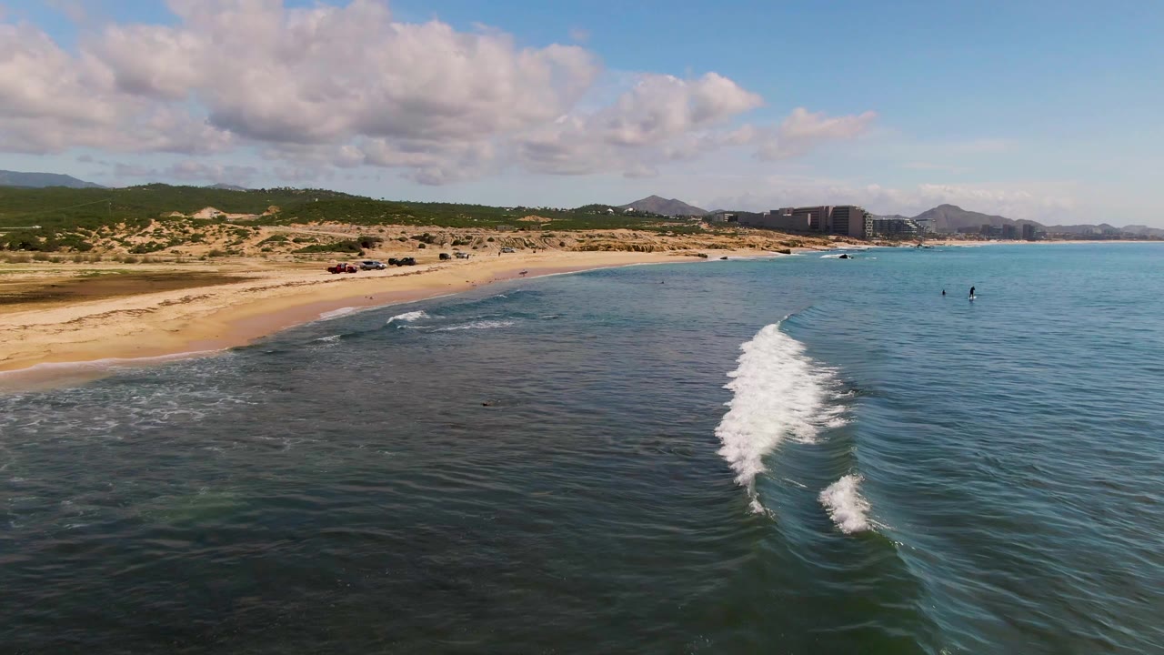 Aerial frame from El Tule Los Cabos showing the wave beginning to break across shallow blue water beside tan sand, dune vegetation, distant hills, and no visible people.