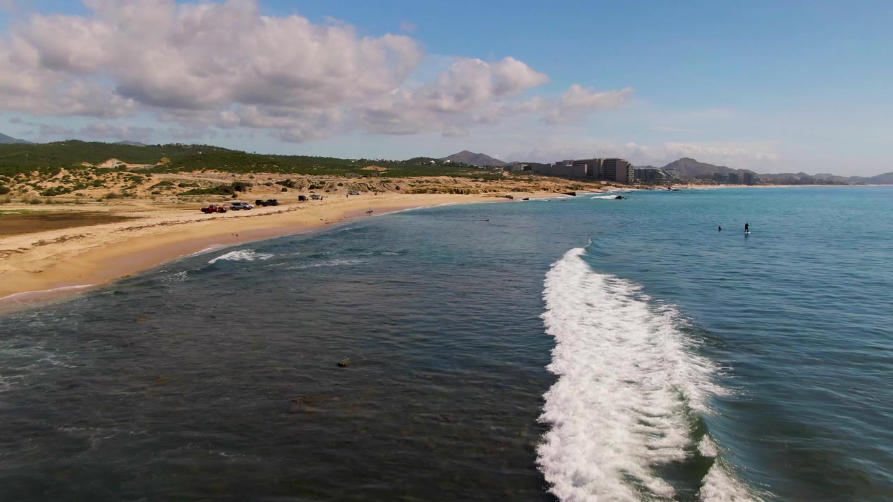 El Tule Los Cabos aerial view with a wider white surf line crossing the right side of the frame, clear blue water, sandy beach, low dunes, mountains, clouds, and no visible people.