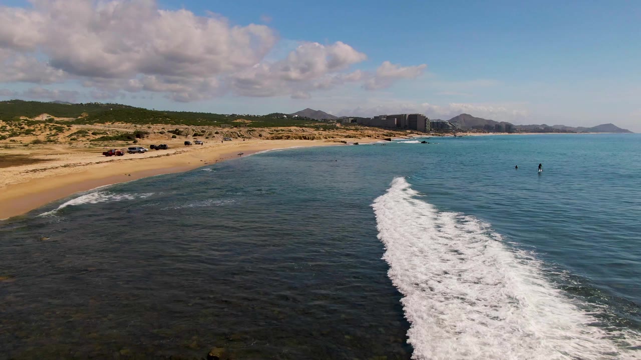 Aerial view of surf curling toward shore with dunes and hills beyond