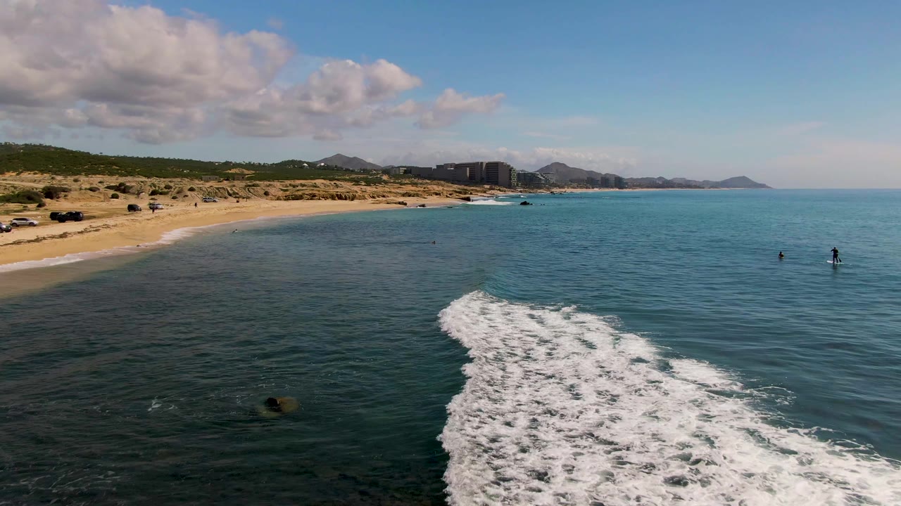 At El Tule Los Cabos, a white foamy wave spreads across shallow blue-green water near a sandy beach, with swimmers offshore, rocks at the left, distant buildings, hills, and clouds.