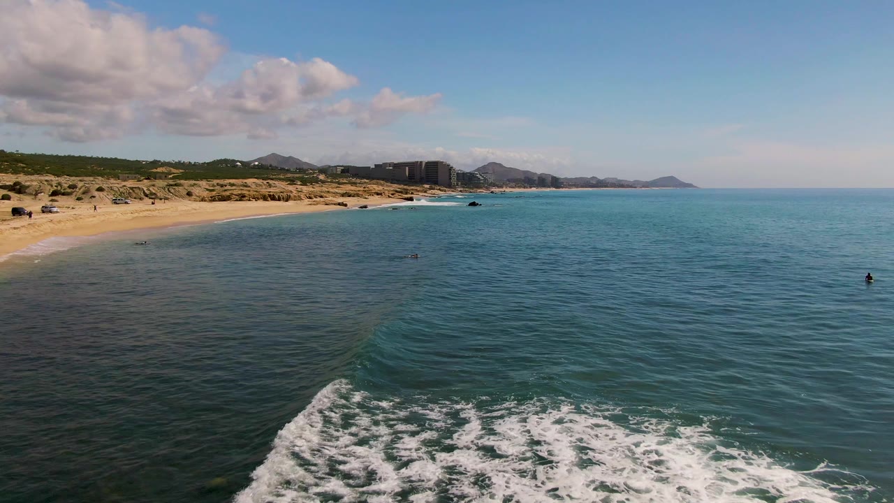 At El Tule Los Cabos, receding surf foam crosses the lower foreground, with the sandy beach to the left, small swimmers in the water, rocks near the shoreline, distant buildings, hills, and a bright sky.