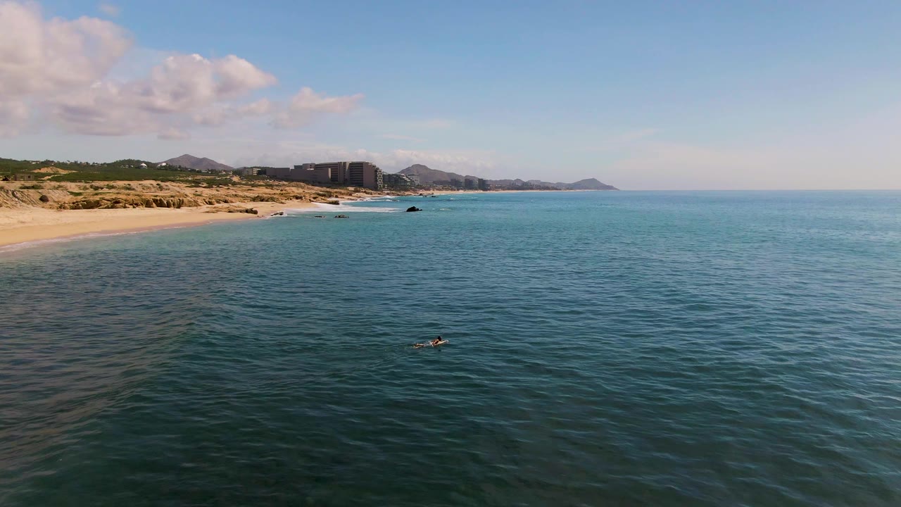 Opening aerial frame over El Tule Los Cabos shows blue-green ocean, sandy beach at left, distant buildings, low hills, and a small swimmer-like figure in the water.