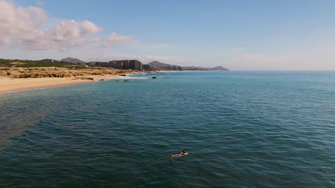 At 0:01, the El Tule Los Cabos aerial view holds more open ocean in the foreground while the sandy beach and distant buildings remain along the left background.