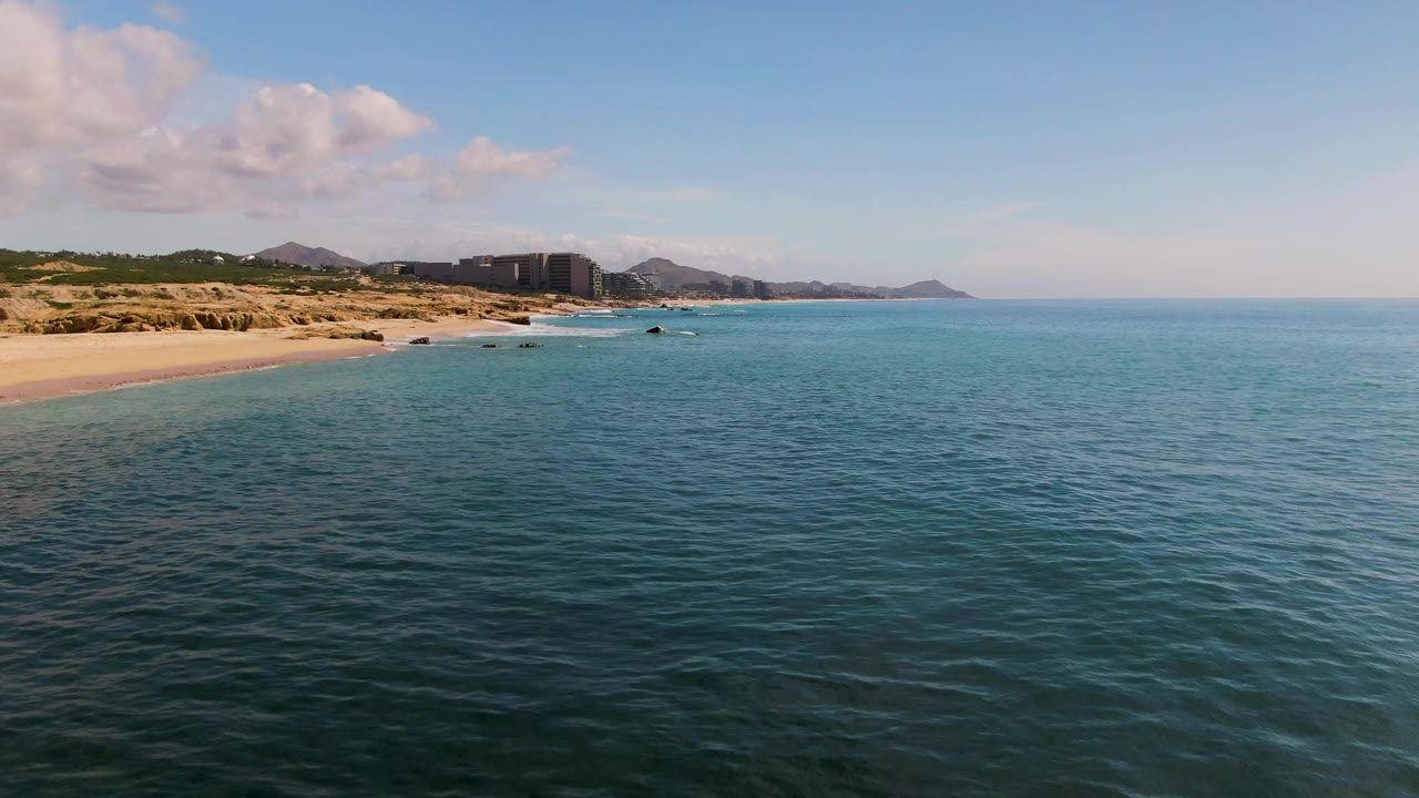 At 0:02, the swimmer is no longer apparent, leaving El Tule Los Cabos ocean, sandy shore, distant buildings, hills, and a partly cloudy sky.