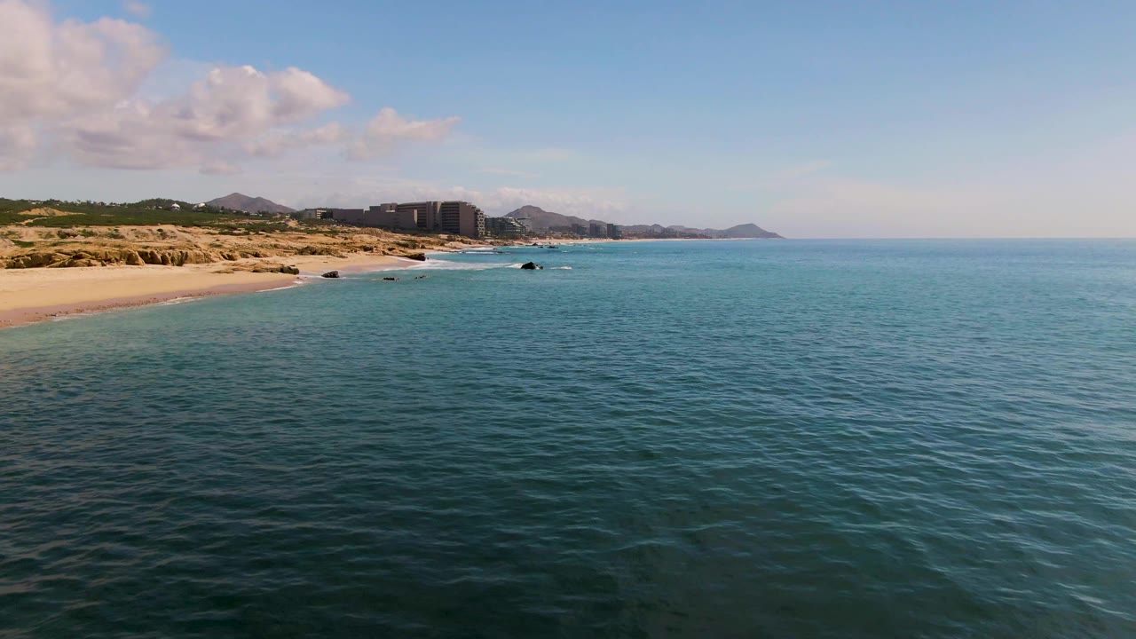 Aerial coastline view with turquoise water, sandy beach, rocks, buildings, and hills.