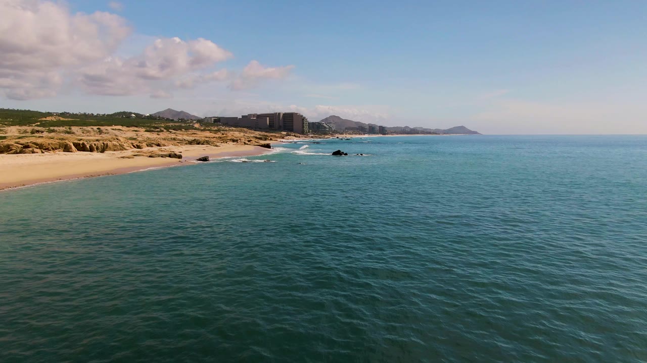 Elevated view of blue-green ocean beside a sandy beach with rocks and distant buildings.