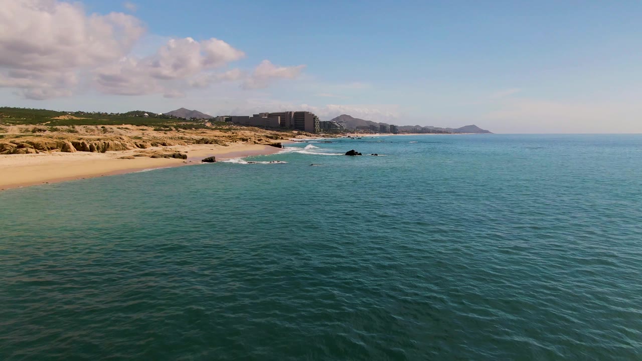 At 0:01 in El Tule Los Cabos, small surf lines reach the sand and rocks as the elevated view holds the ocean foreground, dune-backed beach, distant buildings, hills, and no visible people.
